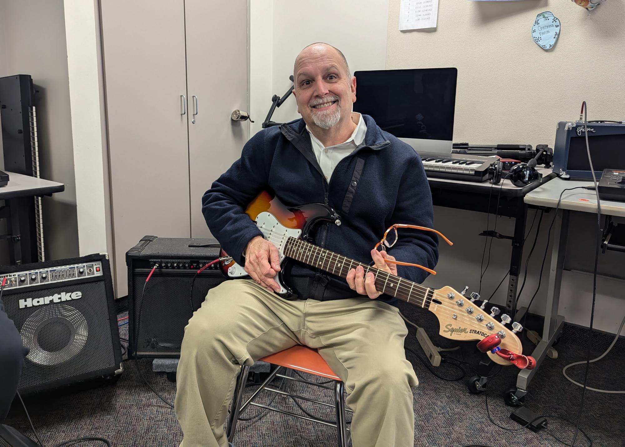 Comprehensive Music Technology teacher, Mr. Daniel Tumolo, smiles while showing off his guitar in his K032 classroom